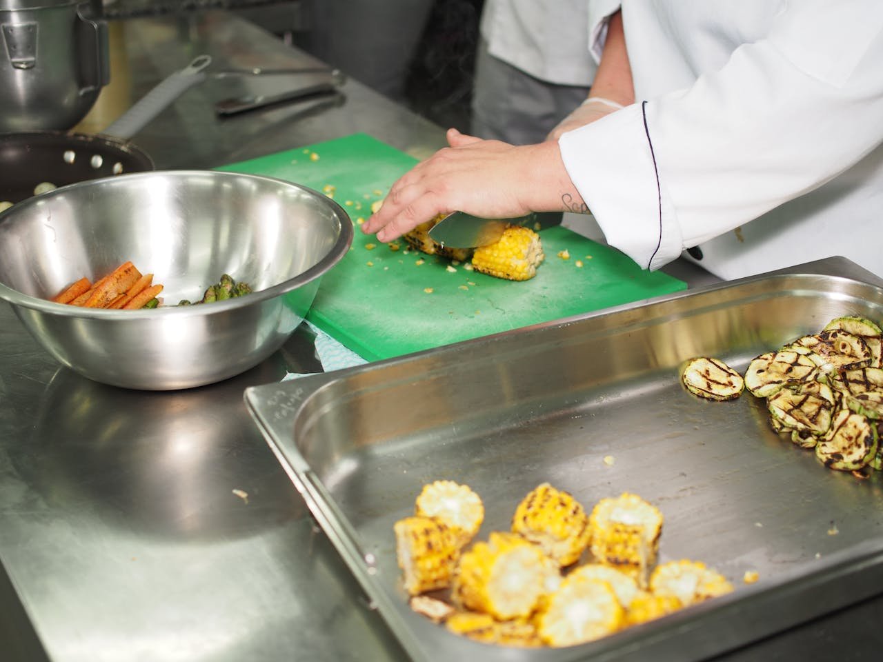Chef slicing grilled corn and zucchini in a professional kitchen setting.